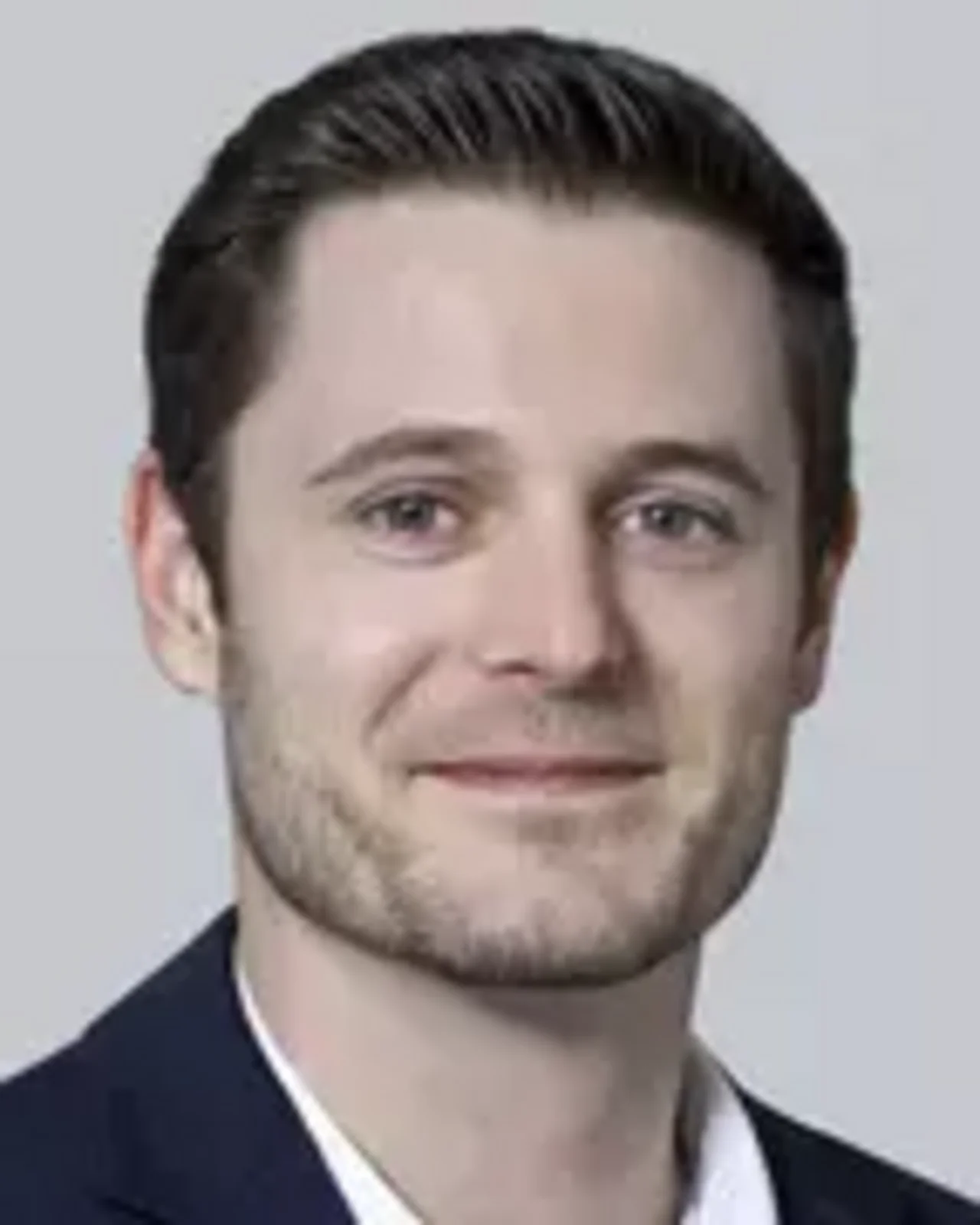 Porträt eines Mannes mit kurzem, dunkelbraunem Haar und leichtem Bart. Er trägt einen dunklen Anzug mit einem weißen Hemd und hat ein freundliches Lächeln. Der Hintergrund ist neutral und hell.