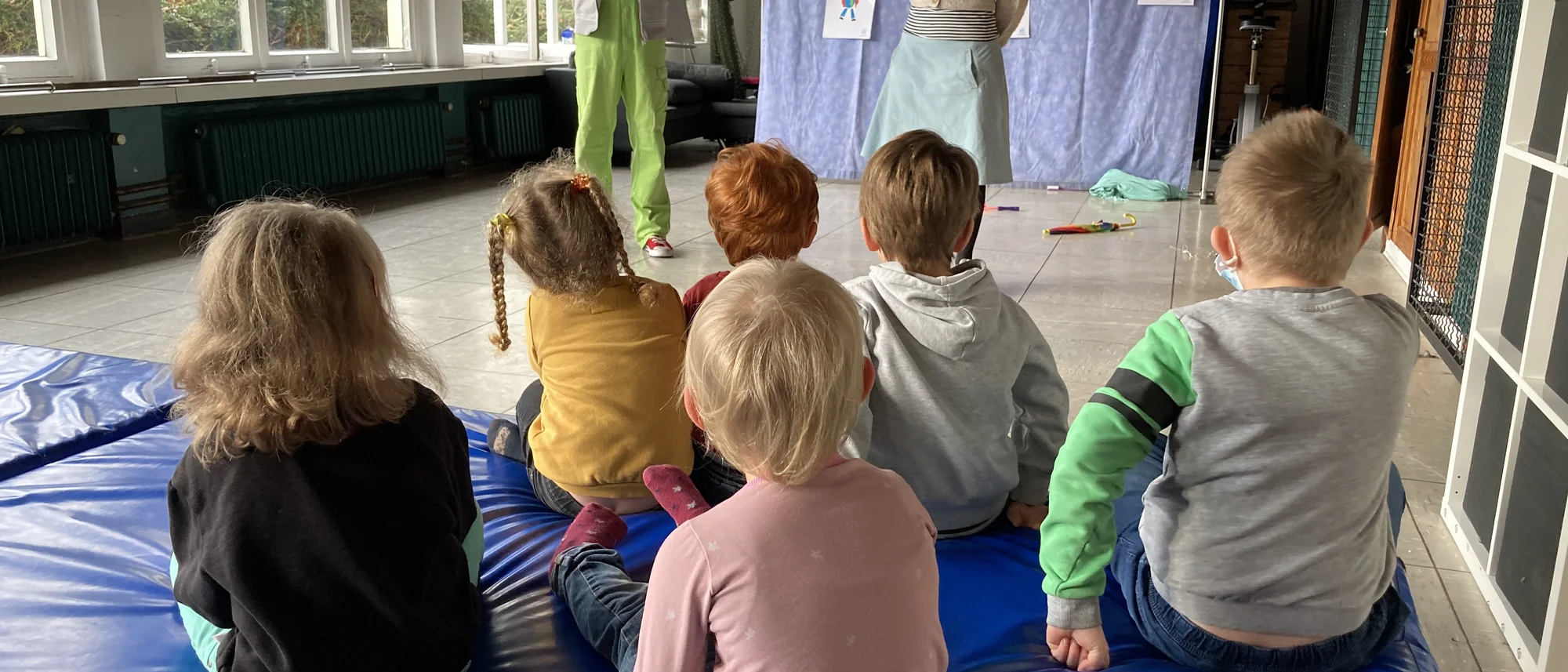 Mehrere Kinder sitzen auf blauen Matten und schauen auf eine improvisierte Bühne, vor der zwei Erwachsene stehen. Eine Frau mit Maske und heller Kleidung spricht zu den Kindern, während ein Mann in grüner Latzhose im Hintergrund mit einer Kamera eine Szene aufnimmt. An einem blauen Vorhang hinter ihnen hängen bunte Kinderzeichnungen. Der Raum ist hell, mit großen Fenstern und Heizkörpern entlang der Wand.