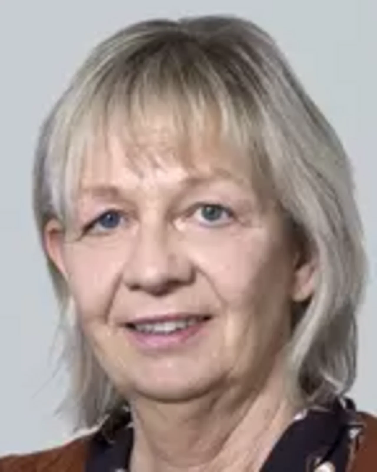 Porträt einer Frau mit schulterlangem, blond-grauem Haar und freundlichem Ausdruck. Sie trägt eine braune Jacke über einem dunklen Oberteil. Der Hintergrund ist neutral und hell.